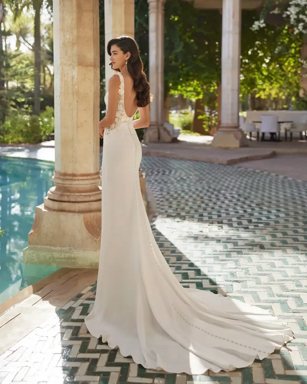 A woman in a sleek white gown stands by a pool, facing away, with her hair styled in loose waves. The setting features elegant columns and lush greenery.