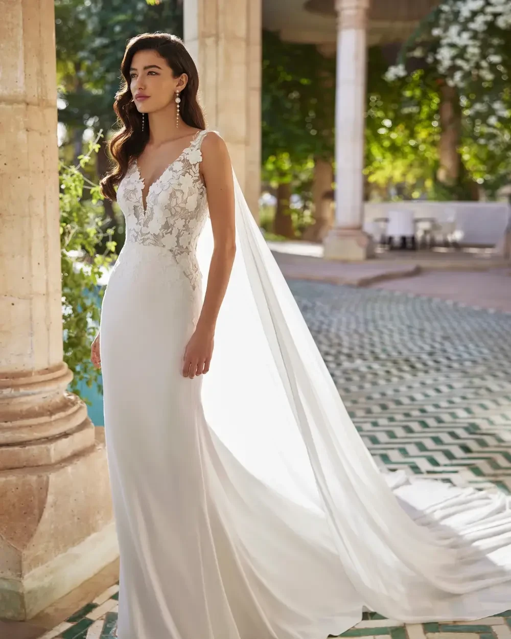 A woman in a sleek white gown stands by a pool, facing away, with her hair styled in loose waves. The setting features elegant columns and lush greenery.