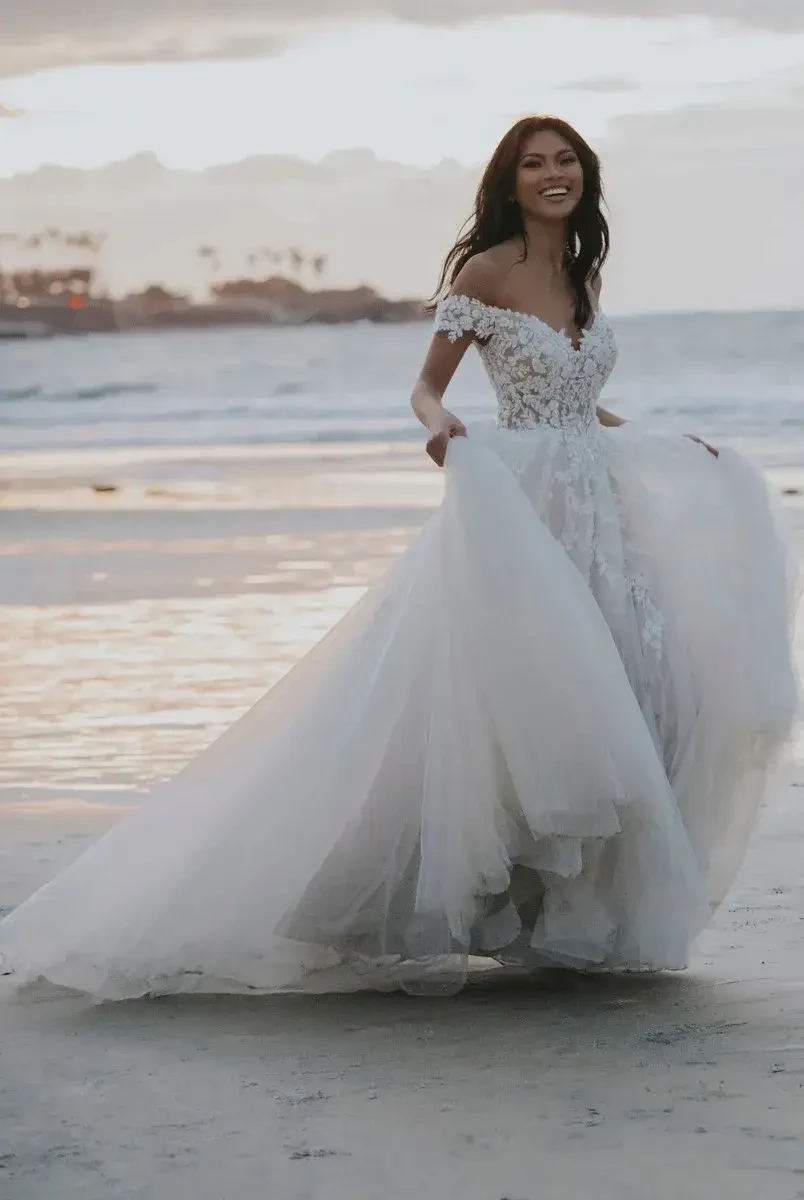 A woman in a flowing white gown stands on a beach, smiling and enjoying the ocean breeze.