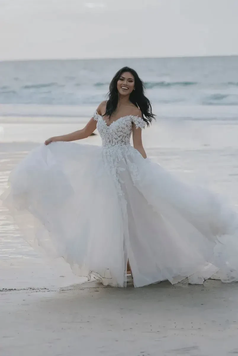 A woman in a flowing white gown stands on a beach, smiling and enjoying the ocean breeze.