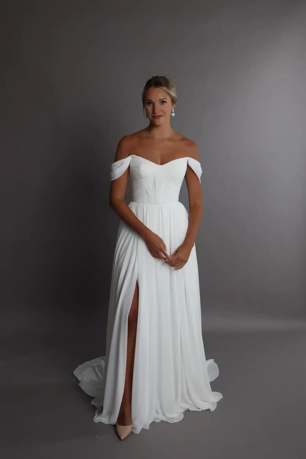 A woman in a white off-the-shoulder wedding dress with a thigh-high slit, standing against a gray background.