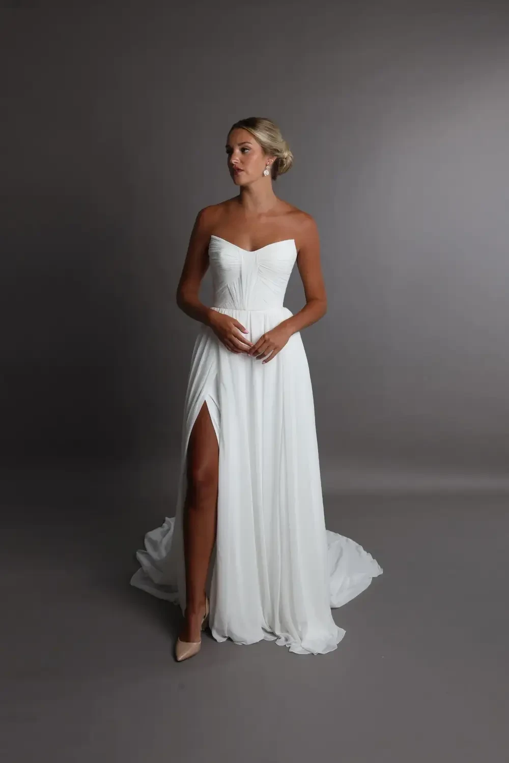 A woman in a white off-the-shoulder wedding dress with a thigh-high slit, standing against a gray background.