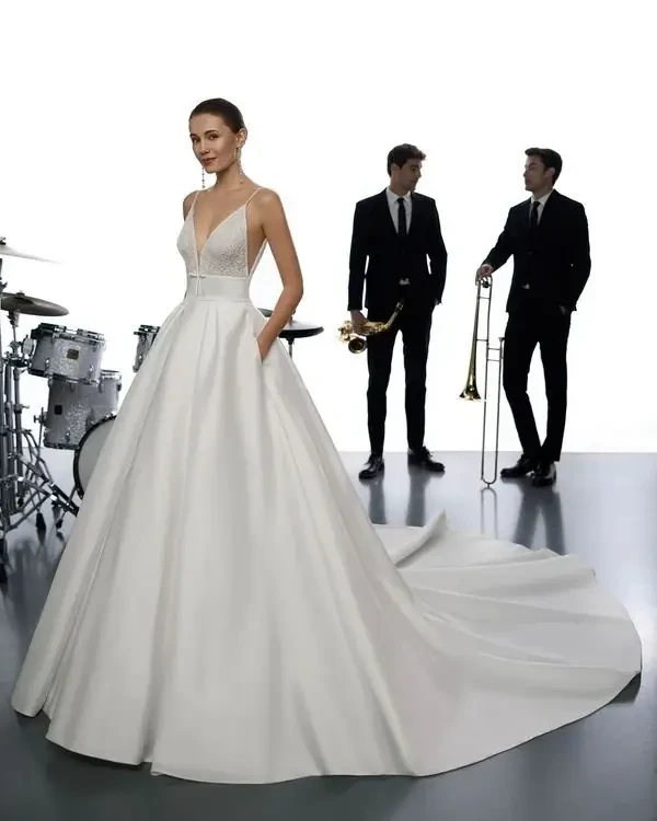 A model wearing a white wedding dress with a full skirt and a fitted bodice stands in front of a band consisting of two musicians dressed in black suits, one holding a saxophone and the other a trombone, with a drum set in the background.